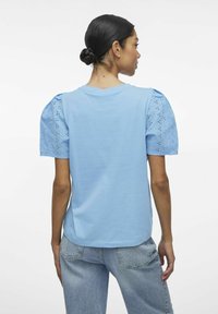 Jasnoniebieska koszulka z krótkim rękawem. Tył ma bufiaste rękawy wykonane z ażurowej tkaniny z wyciętymi wzorami kwiatowymi. Standardowy dół koszulki.