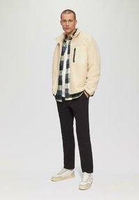 Beige fleecejack met ritszak op de borst, zwart-groene geruite shirt, zwarte broek en witte sneakers met beige accenten.