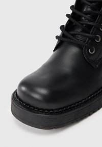 Bota de cuero negro con punta redondeada, que presenta una superficie texturizada, detalles trenzados en la suela y cordones negros pasados por ojales de metal.