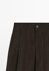 Tmavě hnědé kalhoty s vzhledem semiše s postranními kapsami, poutky na pásek a předním zapínáním na knoflík na bílém pozadí.
