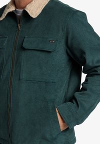 Chaquetón de pana en color verde azulado con un cuello de sherpa color crema, dos bolsillos en el pecho y bolsillos laterales. Cuenta con un cierre metálico y textura acanalada.