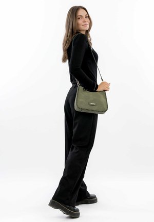 Sac en cuir vert porté à l'épaule, avec des détails cousus et une plaque logo en métal, porté avec un haut à manches longues noir et un pantalon sur fond blanc.