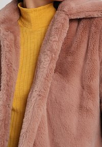 Faux fur-kappa i mjuk rosa med en plyschtextur; har en avspänd passform och en bred krage. Bärs över en gul ribbad tunn hals.