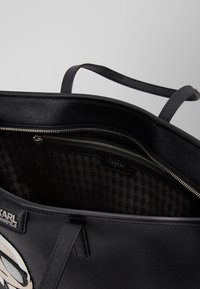 Zwarte totebag met een ritsluiting, gestructureerde buitenkant en een houndstoothpatroon aan de binnenkant. Inclusief dubbele handvatten en logo-detail.