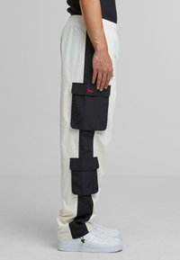White cargo pants with black side panels, multiple large pockets, and elastic waistband. Paired with white sneakers.