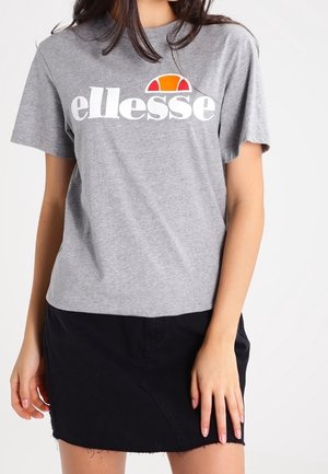 Femme portant un t-shirt gris ellesse avec un logo orange et une jupe en denim noire, debout devant un fond uni.