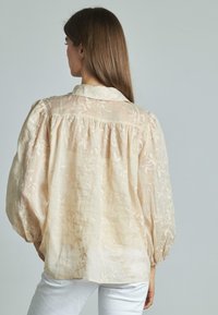 Blusa beige chiaro con ricami floreali, maniche a sbuffo lunghe e colletto. Il tessuto sembra trasparente e testurizzato, con dettaglio arricciato sul retro.