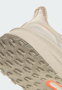 Béžová bežecká obuv s textúrovanou podrážkou, zobrazuje ružový akcent na päte a reflexné časti. Hladký materiál vrchnej časti.