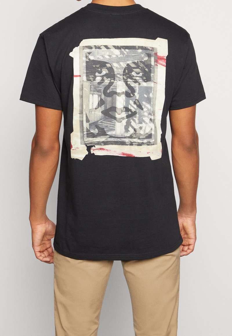 Svart bomullst-shirt med en grafisk tryck på baksidan som visar ett ansikte i en ram, omgiven av beige tejp och röda accenter.