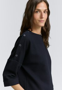 Marineblauw getextureerd top met driekwartmouwen, voorzien van decoratieve knopen op de schouders en een ronde halslijn.