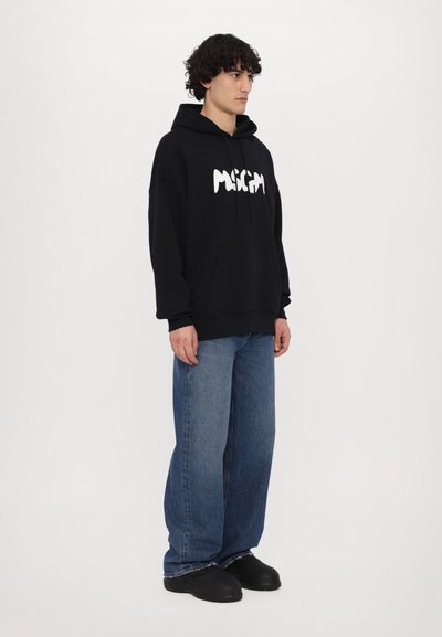 Felpa nera con logo bianco, tasca frontale, coulisse, abbinata a jeans blu a gamba larga e scarpe slip-on nere. Sfondo semplice.