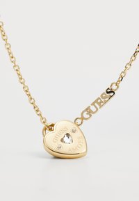 Gold heart-shaped pendant necklace with engraved “GUESS AMORE,” clear stone accents, and linked chain featuring the brand name "GUESS."