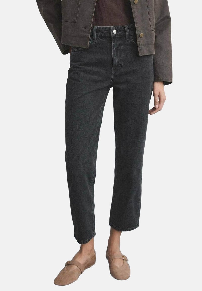 Personne portant un jean noir court, une veste marron et des chaussures plates beiges en daim avec des brides à boucle, debout sur un fond blanc.