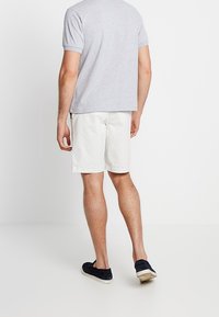 Hombre vistiendo una camisa de manga corta gris claro, pantalones cortos blancos y zapatos negro de goma, de pie con la espalda hacia el frente contra una pared blanca.