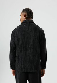Zwart corduroy overhemd met een verticale geribbelde textuur, een kraag met knopen en lange mouwen. Heeft een ontspannen silhouet, gedragen met donkere broek.
