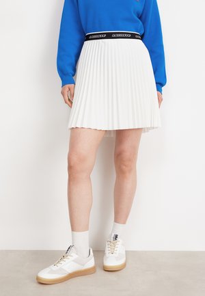 Jupe plissée blanche avec une taille élastique noire arborant "LACOSTE". Assortie à des baskets blanches et des chaussettes blanches côtelées.