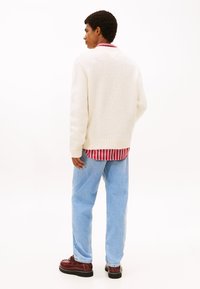 Λευκό πλεκτό πουλόβερ με ριμπ μανσέτες, φορεμένο πάνω από κόκκινο και λευκό ριγέ πουκάμισο, σε συνδυασμό με ανοιχτό μπλε τζιν και καφέ παπούτσια με κορδόνια.