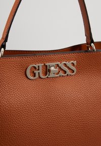 Sac à main en cuir marron texturé avec logo GUESS métallique et deux bandoulières sur un fond clair uni.