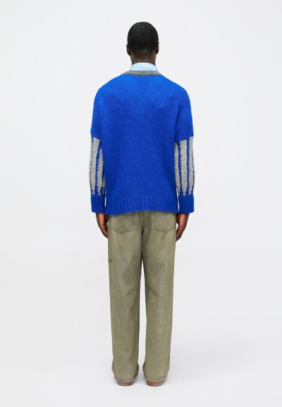 Μπλε πλεκτή μπλούζα με γκρι και εκρού ρίγες στα μανίκια, συνδυασμένη με ανοιχτό πράσινο παντελόνι. Η υφή φαίνεται απαλή, με χαλαρή εφαρμογή.