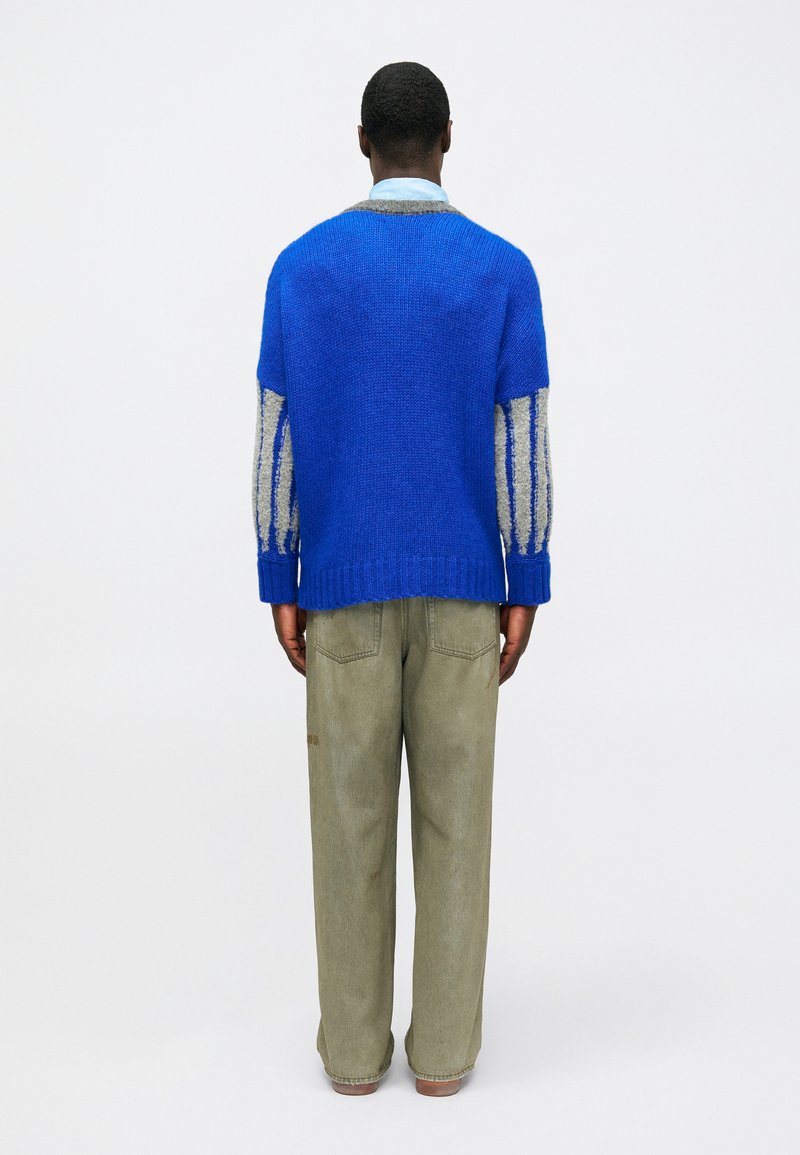 Jersey de punto azul con mangas a rayas en gris y blanco roto, combinado con pantalones verde claro. La textura parece suave y tiene un diseño de corte holgado.