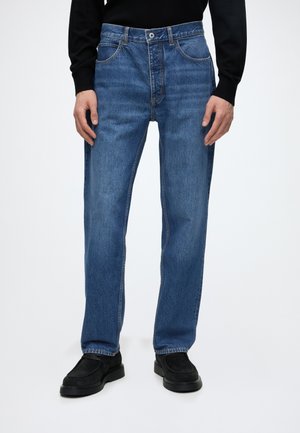 Persoon die blauwe rechte jeans met zichtbare stiksels en zwarte schoenen draagt, staand tegen een lichtgrijze achtergrond.