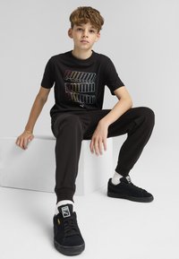Zwart T-shirt met kleurrijke "PUMA" tekst, zwarte sweatpants en zwarte sneakers. Model zittend op een witte kubus met een neutrale achtergrond.