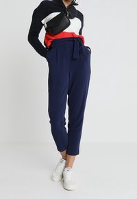 Marineblauwe hoge taille broek met een strikceintuur, voorzien van een taps toelopend ontwerp. Combineert met een kleurgebroken trui en witte sneakers.