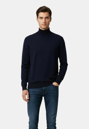 Uomo in piedi che indossa un maglione nero a collo alto e jeans blu con una mano in tasca su uno sfondo chiaro uniforme.