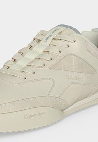 Baskets beige Calvin Klein avec des panneaux texturés, des lacets en tissu et le nom de la marque visible sur le côté, la languette et la semelle.
