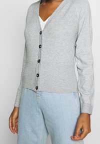 Cardigan grigio a coste con scollo a V, dotato di sei bottoni neri lungo il davanti e maniche lunghe aderenti. Indossato sopra una camicia bianca e jeans blu.