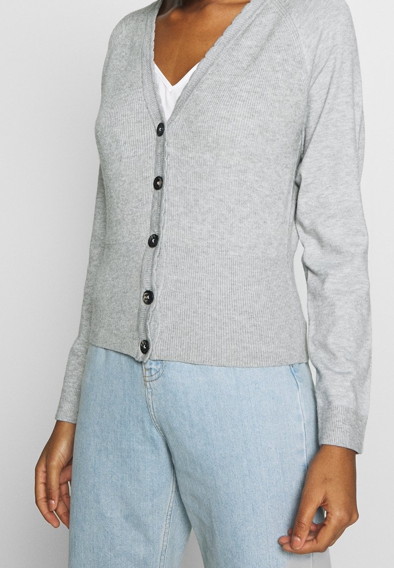Cardigan grigio a coste con scollo a V, dotato di sei bottoni neri lungo il davanti e maniche lunghe aderenti. Indossato sopra una camicia bianca e jeans blu.