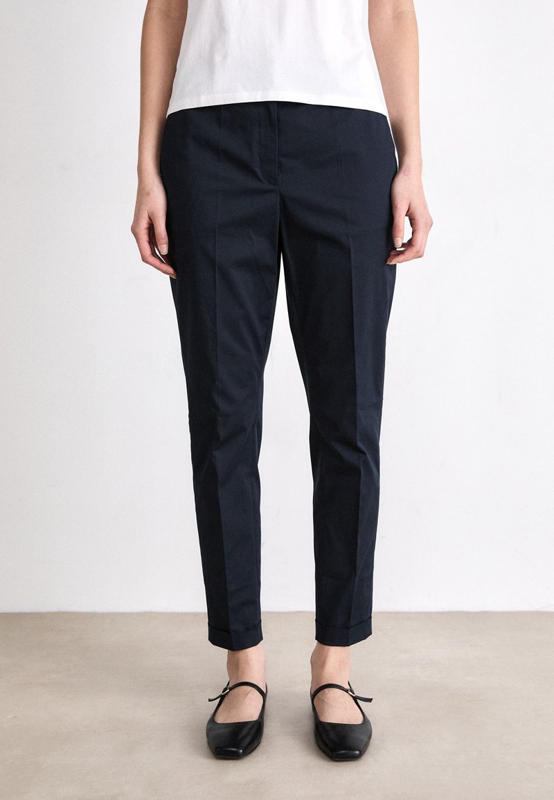 Navy blue tailored pants with front pleats, slim fit, cropped length, and a smooth texture, paired with black flats.