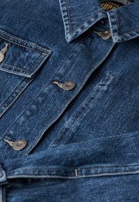 Camisa de mezclilla en azul oscuro con costuras visibles, botones de metal en los bolsillos y el cuello, y sutiles detalles desgastados a lo largo de la tela.