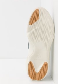 Semelle de chaussure présentant une base en caoutchouc blanc texturé avec un motif chevrons marron. Un logo Lacoste est embossé près du talon.
