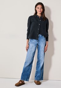 Tmavá, texturovaná košile na knoflíky s dvěma náprsními kapsami, zkombinovaná s volnými světlými modrými džíny a hnědými nazouvacími botami.