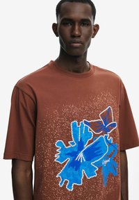 T-shirt en coton marron avec un motif graphique bleu orné de fleurs et d'accents tachetés, col rond et manches courtes.