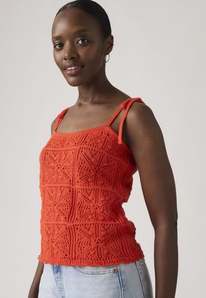 Femme portant un top sans manches rouge en crochet avec des bretelles à nouer et un jean bleu clair, debout devant un fond clair uni.