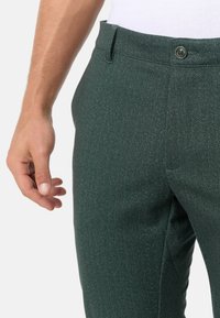 Grüne Fischgrät-Hosen mit strukturiertem Finish, mit Knopfverschluss und seitlichen Taschen, gezeigt von der Taille abwärts, mit einer daneben positionierten Hand.