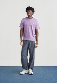 Μωβ βαμβακερό t-shirt με στρογγυλή λαιμόκοψη και μικρό λογότυπο, συνδυασμένο με άνετο σκούρο γκρι παντελόνι και λευκά αθλητικά παπούτσια σε μπλε πάτωμα.