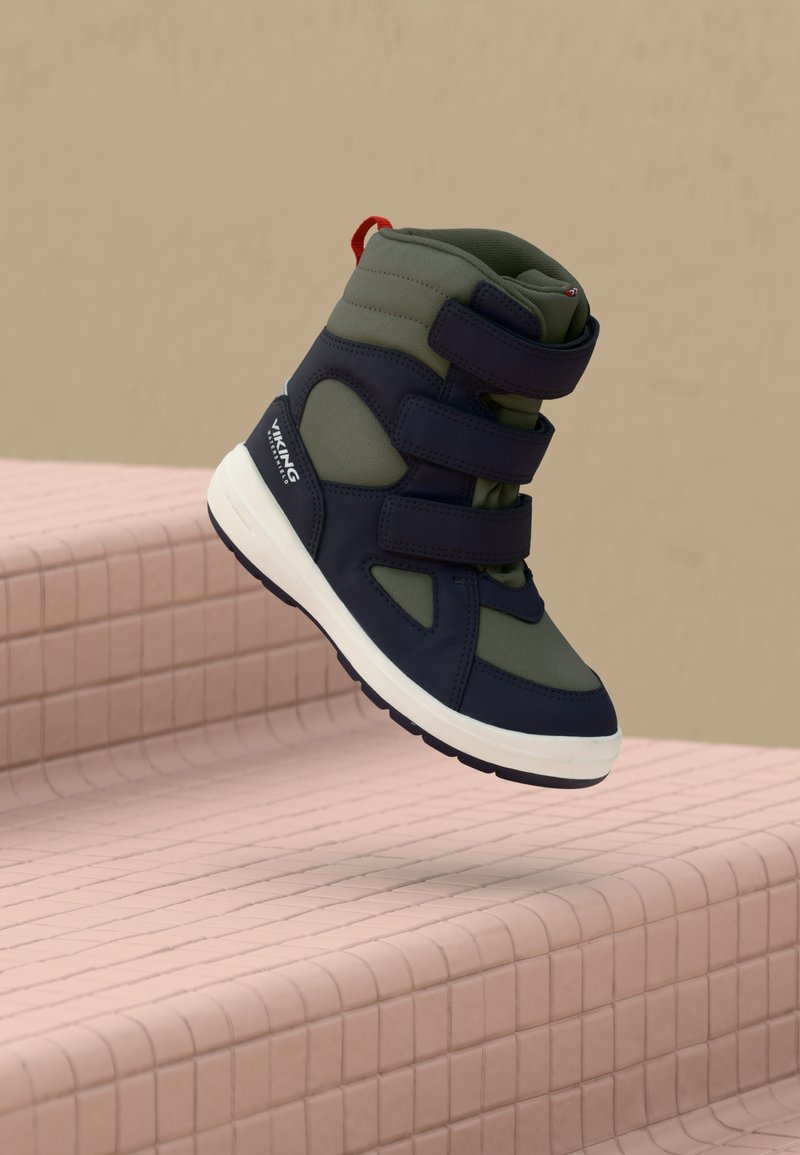 Sapatilhas altas em azul marinho e verde com duplas correias de Velcro e uma alça vermelha. Apresenta uma sola branca com aderência texturizada.