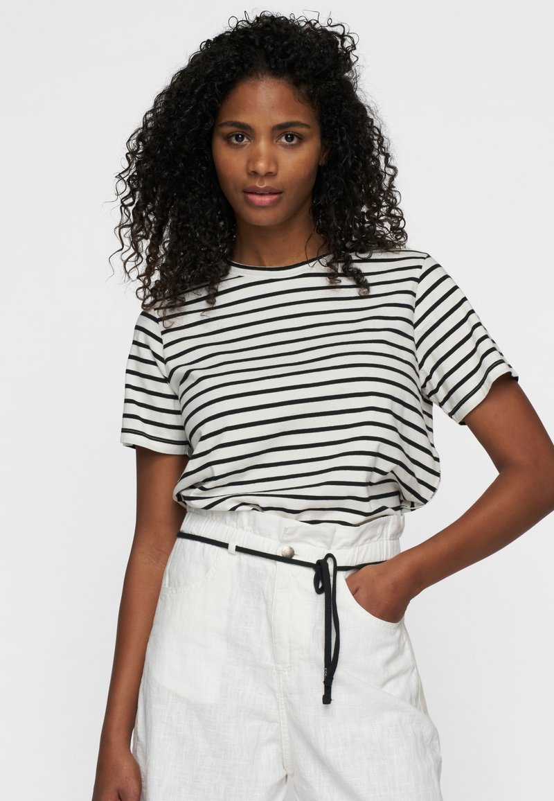Camiseta de manga corta a rayas en blanco y negro, combinada con pantalones de lino blancos de cintura alta, que cuentan con un cordón y bolsillos frontales.