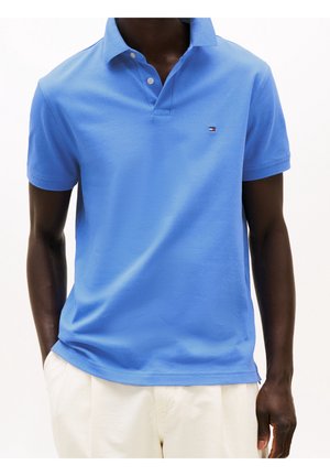 Mężczyzna ubrany w jasno niebieską koszulkę polo z krótkim rękawem i małym logo na piersi oraz białe spodnie, z rękami częściowo w kieszeniach.