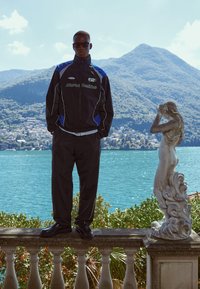 Fekete sportos dzseki kék részletekkel és fehér logóval, hozzá illő nadrággal és fekete cipővel párosítva. A modell egy peremen áll egy szobor mellett.