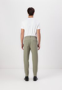 Joggebukser i lys olivengrønn, med elastisk linning, to baklommer og en smal nederdel. Stoffet ser lett og strukturert ut.