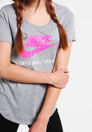 Ung kvinde med rødt flet hår iført en grå Nike International t-shirt med lyserøjt logo, holder venstre arm med højre hånd.