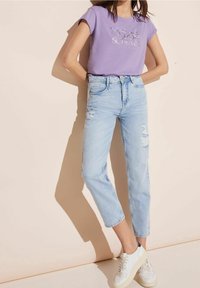 Nainen, jolla on päällään laventelin värinen graafinen t-paita ja vaaleansiniset kuluneet farkut valkoisilla lenkkareilla, seisoo vaaleanbeigeä seinää vasten.