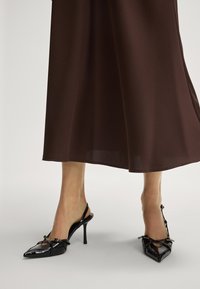 Escarpins à talons aiguilles en cuir verni noir, à bout pointu, avec un talon fin et un détail de strap décoratif, portés avec une jupe fluide marron.