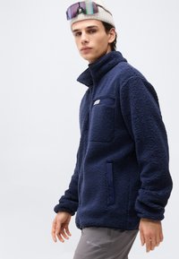 Νέος άνδρας φορώντας μπλε σκούρο φλις μπουφάν, γκρι παντελόνι, μπεζ σκούφο και ανακλαστικά γυαλιά ηλίου στο κεφάλι, μπροστά σε ένα απλό φωτεινό φόντο.