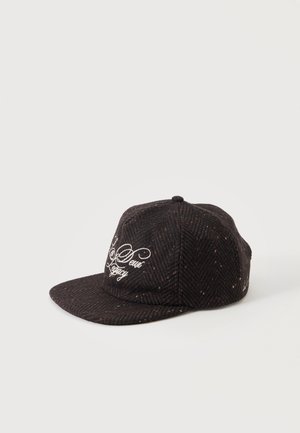 Casquette plate en tweed marron foncé avec broderie blanche en cursive indiquant « "New Legacy" » sur le panneau avant, présentée sur un fond uni.