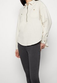 Camisa de color beige claro con cuello y media cremallera, dos bolsillos en el pecho, textura sutil y dobladillo redondeado; combinada con pantalones ajustados de color gris oscuro.